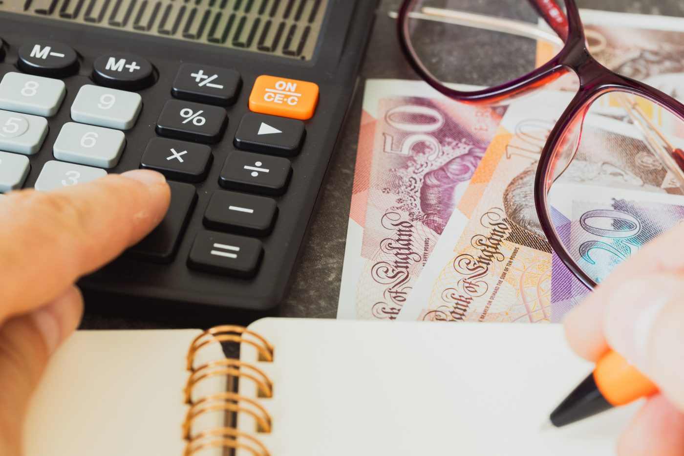 Close-up of calculator, British pound notes, glasses, and notepad.