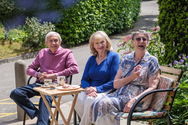 Three seniors enjoy coffee and pastries outdoors.