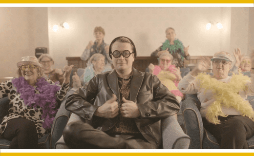 Man in leather jacket with thumbs up, surrounded by older adults in feather boas.