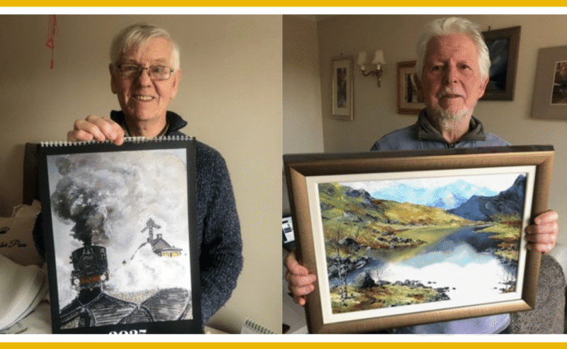 Two men displaying their artwork: a calendar featuring a train and a framed landscape painting.